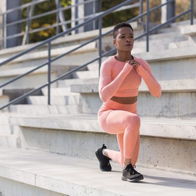 African American woman performs fitness exercises for weight loss, performs squats in a pink suit in the morning in the air near the stadium