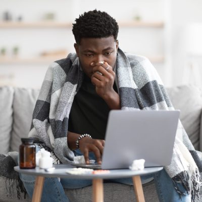 Sick black guy with warm blanket on his shoulders sitting on couch, sneezing and using laptop, looking for coronavirus symptomes on Internet, having video call with doctor, home interior